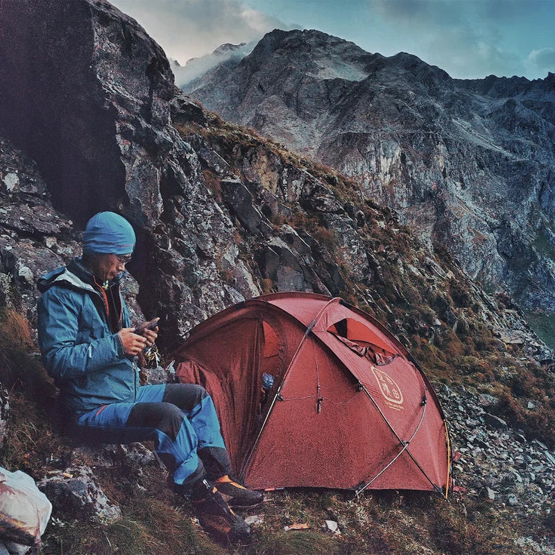 The Free Spirits Whisperwind S red tent on a rocky ridge, a mountaineer resting beside it against dramatic misty peaks.