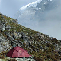 The Free Spirits Whisperwind S red tent pitched on an alpine scree slope, snow-capped mountain rising behind in the mist.