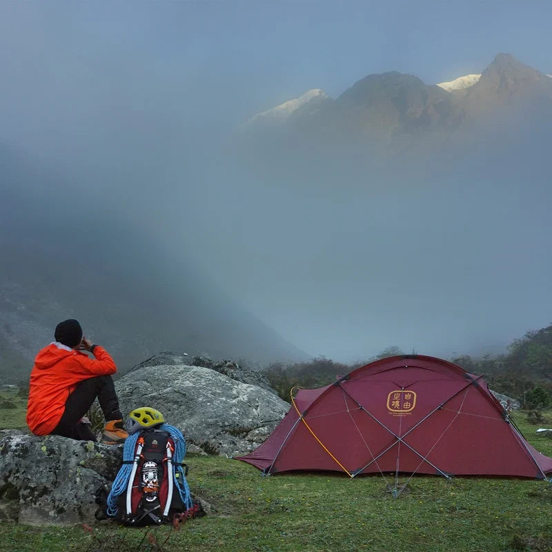 The Free Spirits Whisperwind S red tent on grass, camper with backpack and helmet gazing at misty mountains.