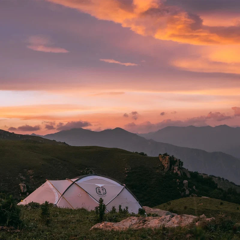 The Free Spirits Whisperwind S tent pitched on a grassy ridge at sunset, warm orange and pink sky over mountains.